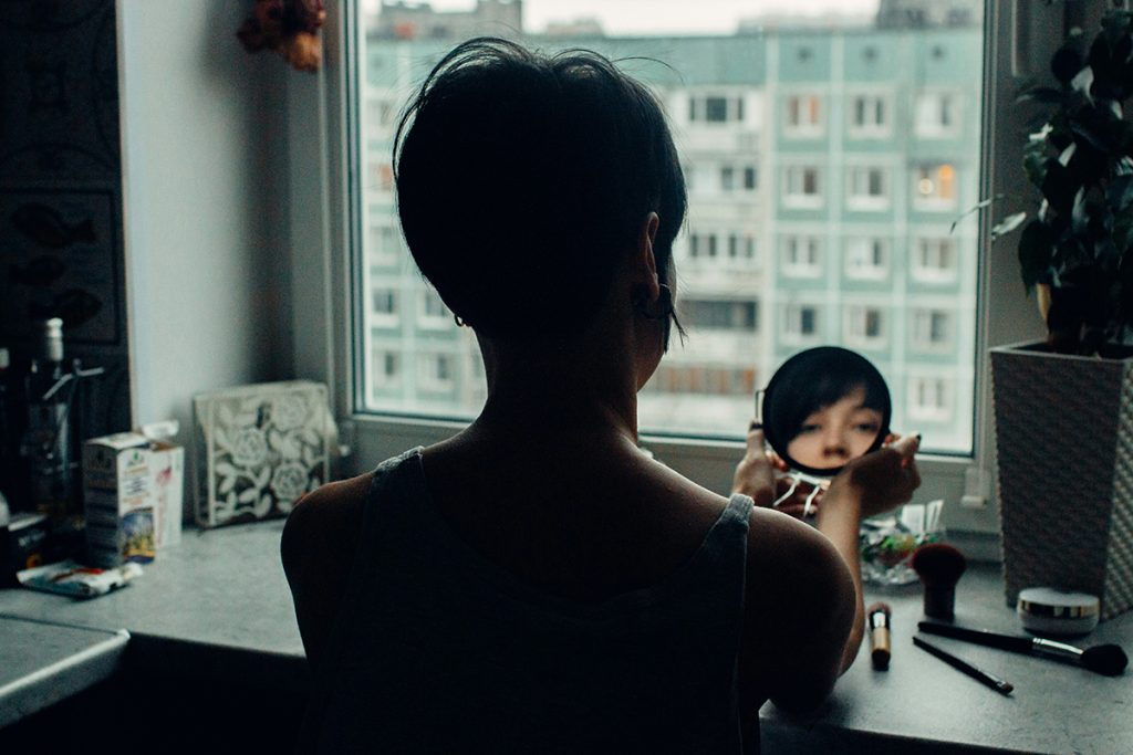 Portrait of a woman seen from the back looking into a small mirror, sitting at a window in a city